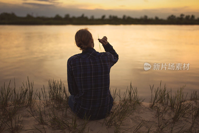 美丽的夏日夕阳背景上的男子剪影。橙色的天空。那家伙正坐在河边。人坐在河岸上