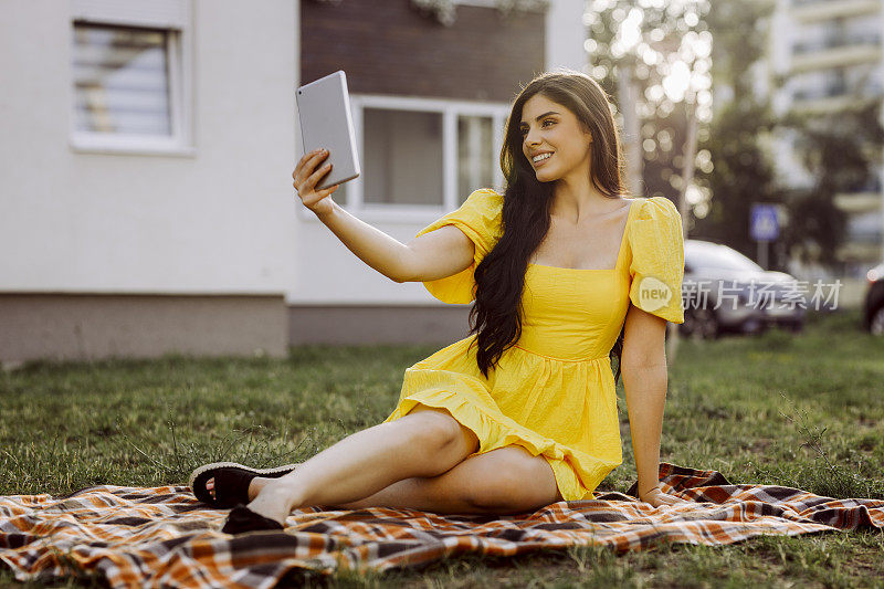 微笑的女人在户外野餐毯子上休息时，正在用数码平板电脑进行视频通话