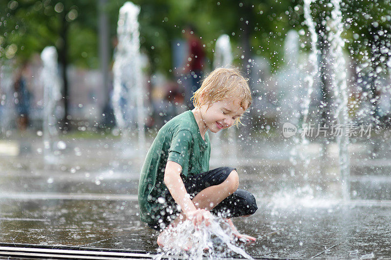 一个阳光明媚的夏日，小男孩在城市喷泉的喷泉之间的广场上玩耍。城市孩子们活跃的夏季休闲活动。