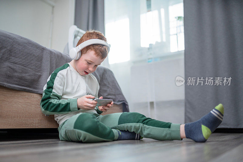 可爱快乐的红发男孩坐在床前的地板上，用智能手机玩游戏