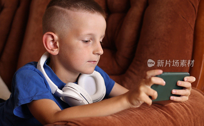 小男孩在家里的沙发上用智能手机玩游戏。孩子用手机玩游戏在线教育社交媒体。小学生正在学习手机游戏。