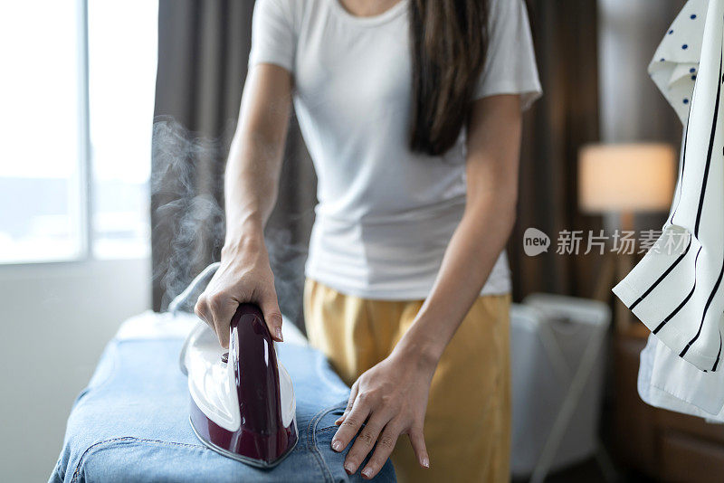 年轻的亚洲成年妇女在家里熨烫她的布，一个快乐的女人在家熨烫她的衣服和微笑的生活方式概念的肖像