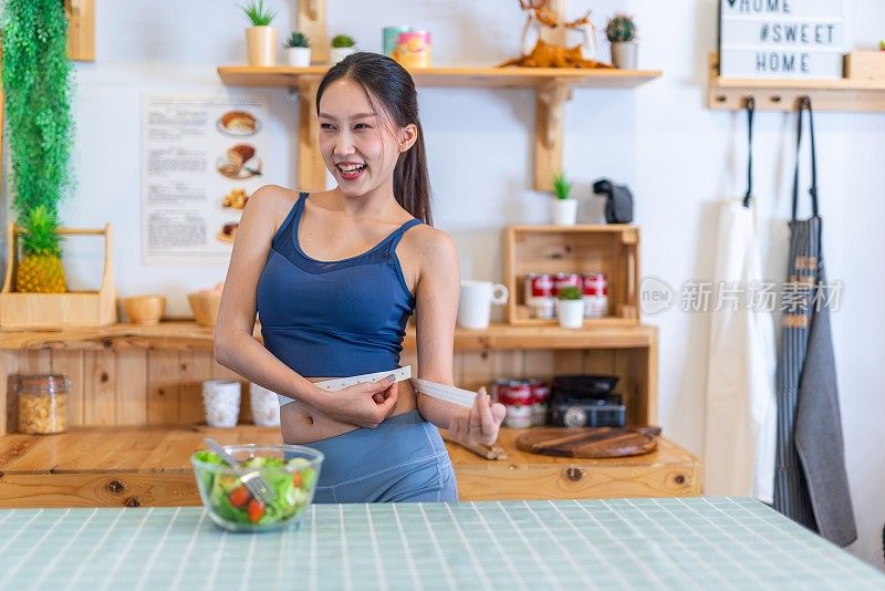 照片的年轻美丽的亚洲妇女女士穿着运动服快乐做腰测量检查她的训练，锻炼和饮食的结果。她家厨房桌上的樱桃番茄沙拉是为了减肥和健康的生活方式