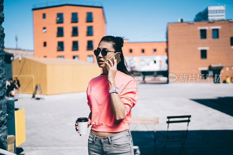 亚洲女学生戴着时髦的太阳镜站在城市街道上，在咖啡休息时间进行国际对话，迷人的千禧女性通过手机应用程序和4g漫游打电话