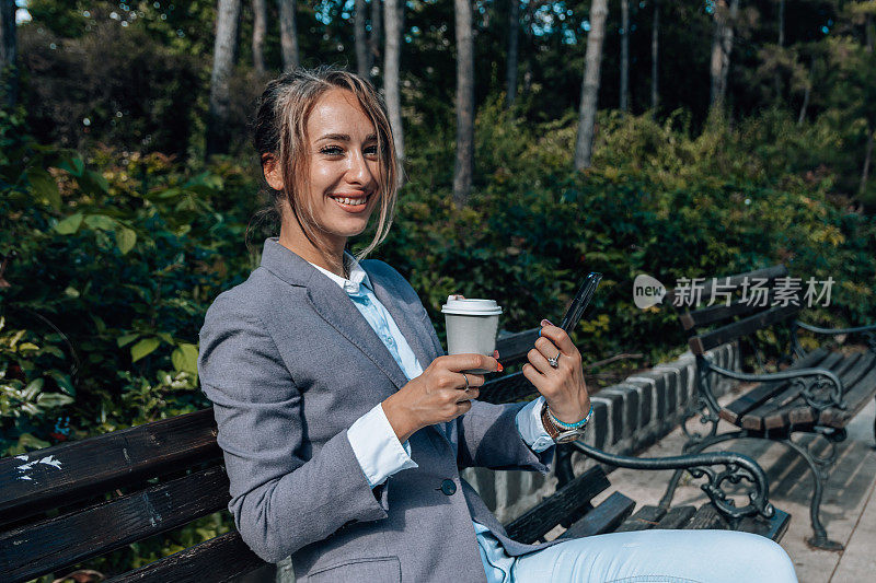 一个女商人坐在长凳上，一边喝着咖啡，一边浏览手机