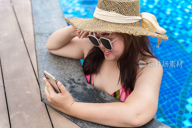 年轻女子橙色swimsuitÂ在游泳池里放松地拿着智能手机。肥胖女人度假旅行用手使用手机