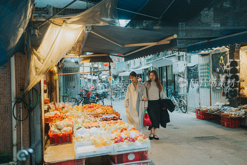 在香港，两名年轻的亚洲女子走过一家商店，店里陈列着各种包装食品，包括海鲜、肉类和零食等干货，这些食品摆放在桌子上，挂在店面上