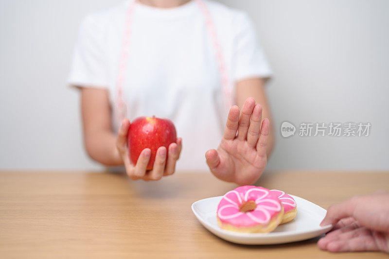女性手持红苹果而拒绝甜甜圈，女性健身在健康水果和不健康的垃圾食品之间做出选择。节食控制，体重减轻，肥胖，饮食方式和营养