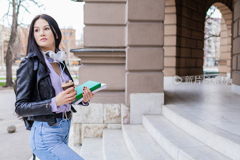 年轻漂亮的女大学生背着双肩包站在大学的楼梯上。她戴着耳机，穿着皮夹克，等待着大学课程的开始。