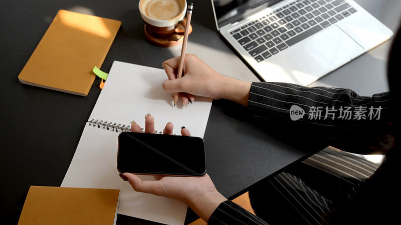 头顶拍摄的女商人在舒适的工作环境中使用智能手机在笔记本上写作
