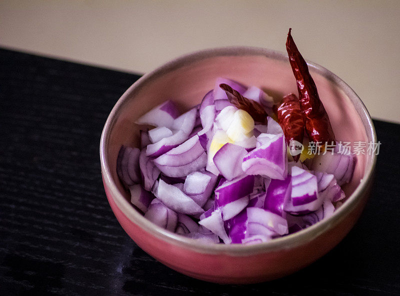在一张黑色的桌子上，把切碎的红洋葱和干泰国辣椒放在一个粉红色的手工花瓶里，背景模糊
