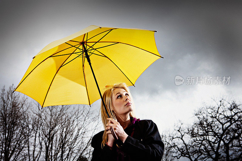 美丽的金发女郎在暴风雨中的黄色雨伞下满怀希望地抬头望着