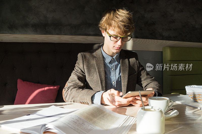 阳光明媚的日子里，年轻的现代男性学生在舒适的餐厅里用平板电脑和书籍工作