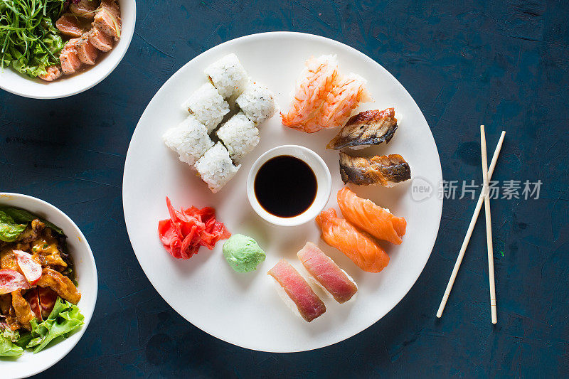 白色圆形托盘上盛有各种寿司卷、生鱼片、山葵，放在深蓝色的木桌竹筷拷贝空间上，顶视图平放