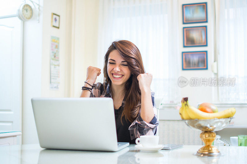 一个快乐的年轻女子在家里的笔记本电脑前举起手臂庆祝成功的肖像。