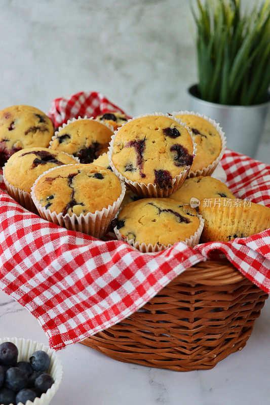 图片:一批自制蓝莓松饼装在纸制蛋糕盒里，柳条篮子里衬着红白格子布，旁边是碗新鲜的蓝莓和咖啡杯，大理石效果的背景，聚焦前景