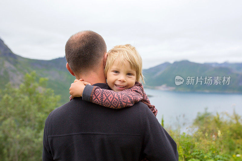 可爱的蹒跚学步的孩子，站在爸爸的肩膀上对着相机微笑，在从罗浮敦到Senja岛的途中，在令人惊叹的挪威自然风光上拍照