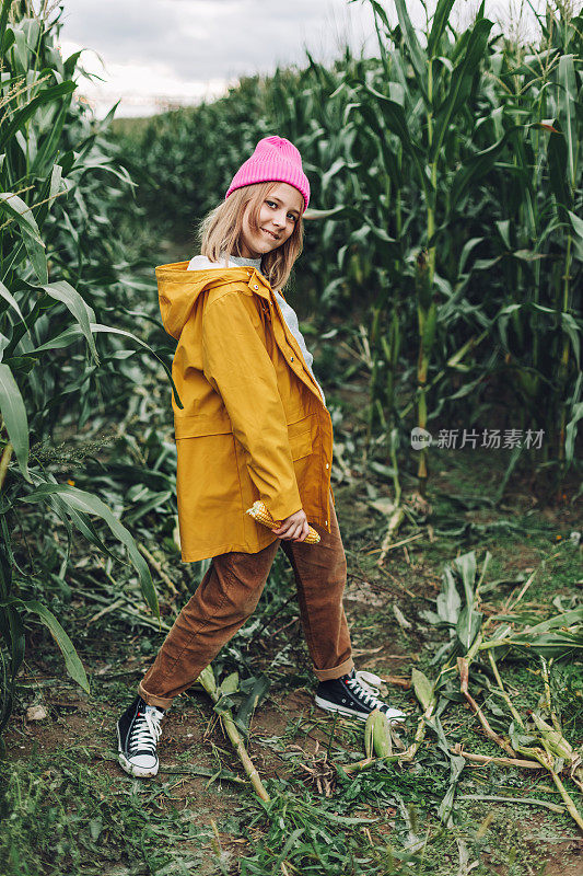 一个穿着黄色雨衣，戴着一顶粉红色帽子的时髦少女在一片玉米地上跳舞和大笑