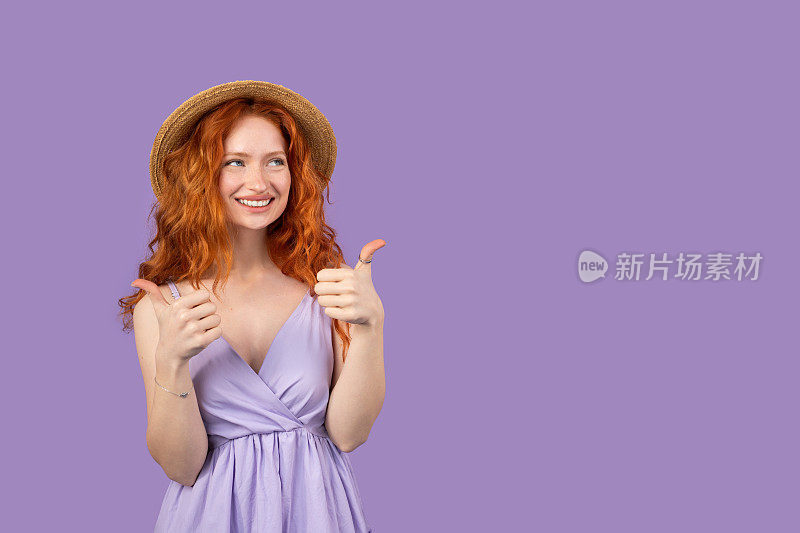迷人的白种人红发年轻女子戴草帽做快乐的拇指手势的手和微笑的姿势在紫色的背景和复制空间。