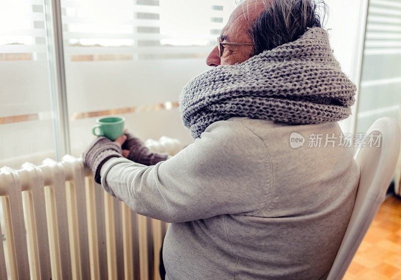 老人在客厅，专注于电加热器。冬天在家里使用加热器。年长的男人在暖手。供暖季节。寒冷的成年男子在家里盖着衣服。