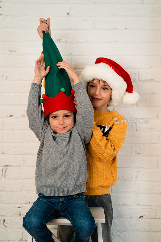 孩子们戴着圣诞老人帽和小精灵帽，在白色的砖墙前摆造型，制作圣诞贺卡