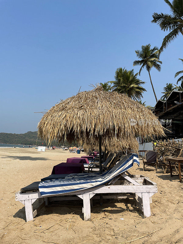 印度果阿，帕洛勒姆海滩，一排木制的蓝色软垫躺椅，在沙滩上晒日光浴，茅草遮阳伞，蓝色和白色条纹的毛巾，边桌