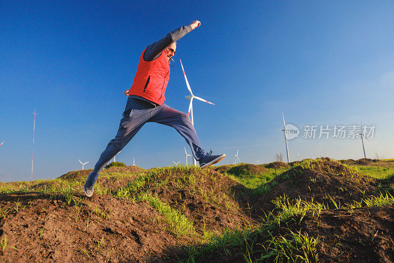 一个高个子男人在风力涡轮机旁的绿色山丘上跳跃