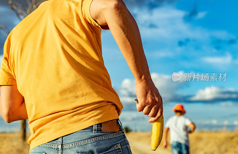 一个无法辨认的男子的后视图，在阳光明媚的乡村环境中打开香蕉套。在模糊的背景中，一个戴着帽子的孩子走开了。