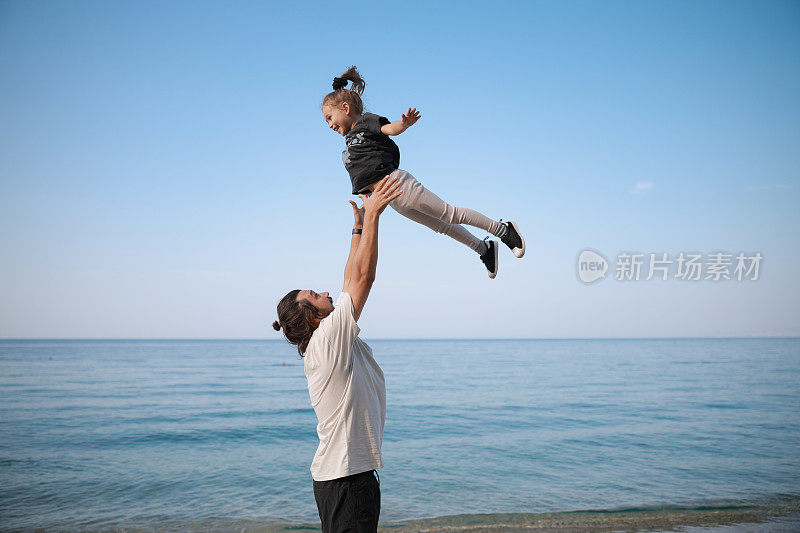 父亲抱着孩子，和家人一起呼吸着空气和蓝天，户外旅行和自由，和女孩和男人一起享受着亲密和海洋。快乐的人，阳光和热带的假期，孩子和爸爸一起飞翔和冒险