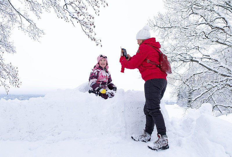 一位年轻的白人妇女用智能手机给一个孩子拍照;孩子坐在一大堆雪里微笑着。
