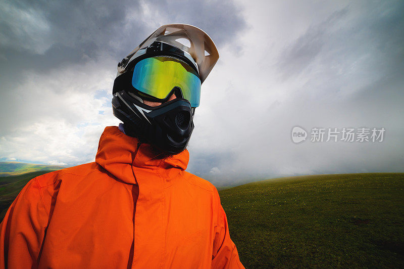 在MTB自行车或摩托车上，骑手戴着头盔和护目镜，全副防护装备站在岩石上，背景是山丘和低云