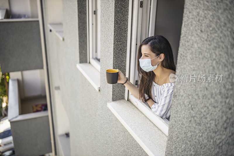 一名年轻的成年女子在因冠状病毒而被隔离期间，倚靠在打开的窗户上，透过窗户看外面，喝着咖啡，戴着医用口罩在家休息