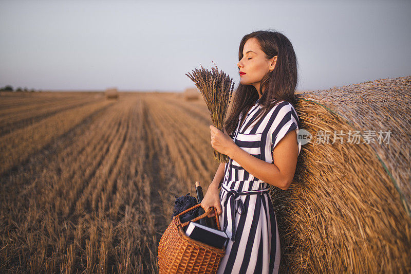 拿着野餐篮的妇女站在收获的田野上，手里拿着草包