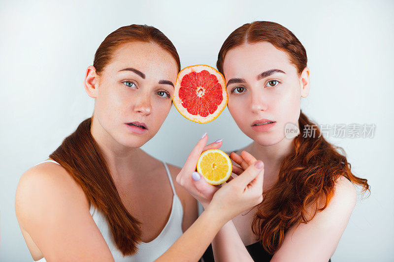两个红头发的年轻女子夹着半个柚子夹在额头之间，手里拿着半个橙子站在孤立的白色背景上，美丽的肖像概念