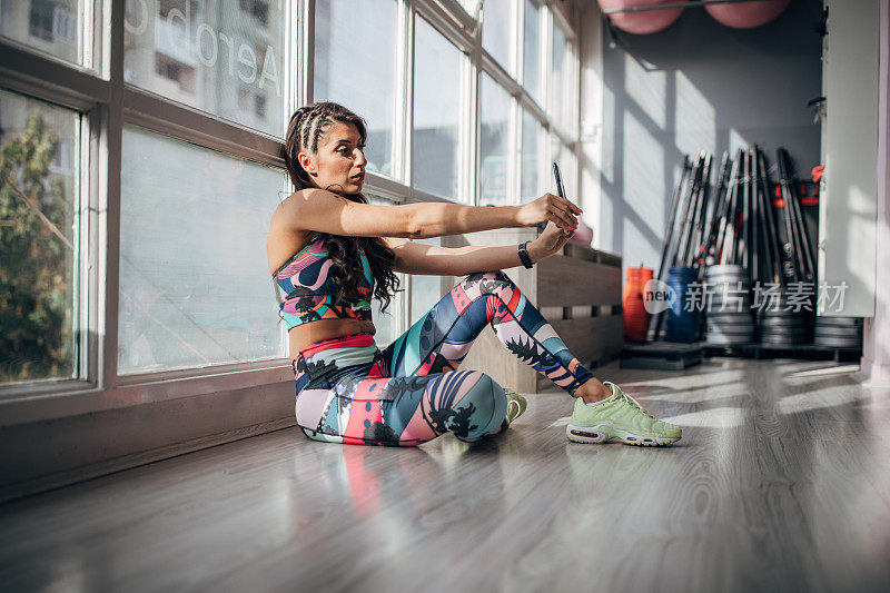 年轻女运动员坐在健身房的地板上用智能手机自拍