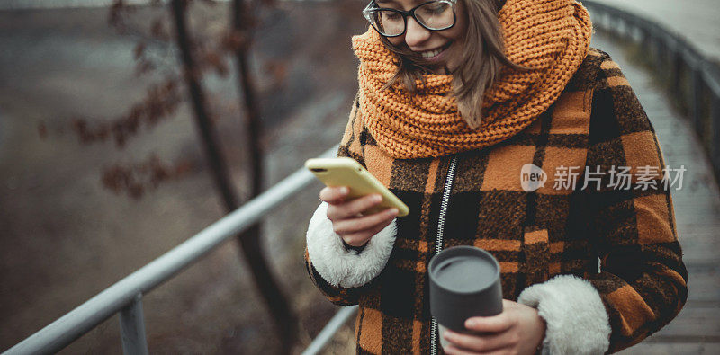 一个年轻的女人站在一个覆盖着秋雾的湖边，走在小路上，喝着她的热咖啡，看着她的手机