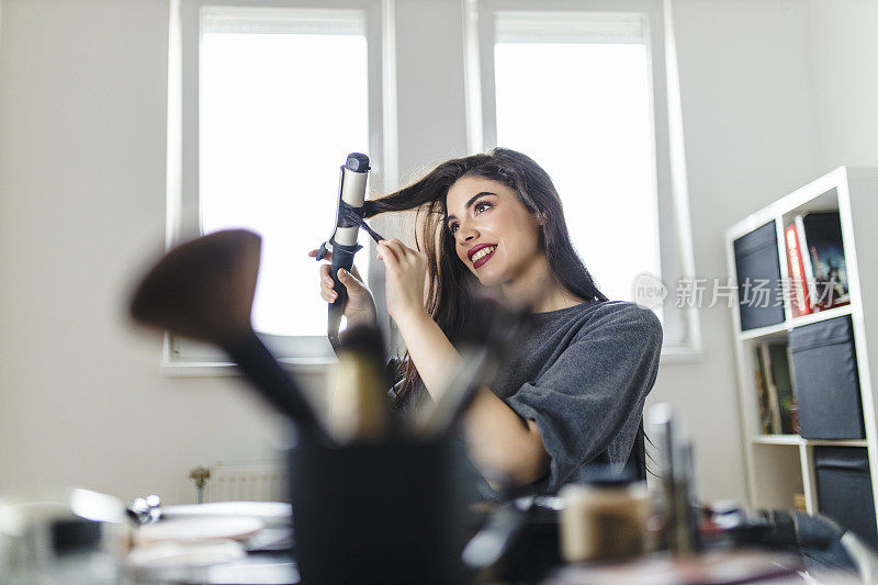 坐在梳妆台边看电脑边用卷发棒的女人。在冠状病毒大流行的时候，一名妇女通过在网上看教程来做头发。