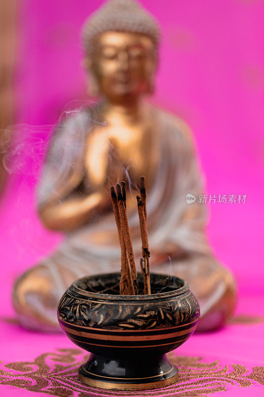 在一个没有聚焦的金佛前面的香碗中冒烟的香和刺绣的粉红色印度教纱丽绳的特写