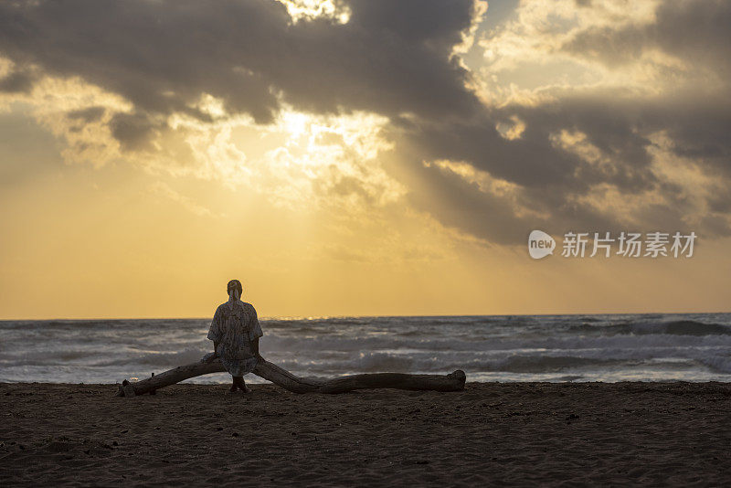 意大利，撒丁岛，夕阳西下，女人坐在沙滩上的树干上看海。