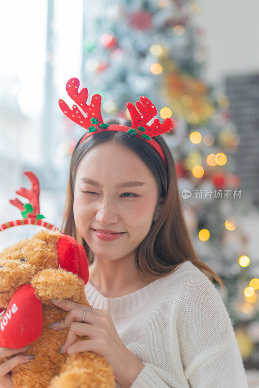 一个年轻美丽友好的亚洲女性女士的肖像照片可爱的圣诞驯鹿发带和柔软的玩具微笑愉快地在一个漂亮的装饰圣诞树的背景前