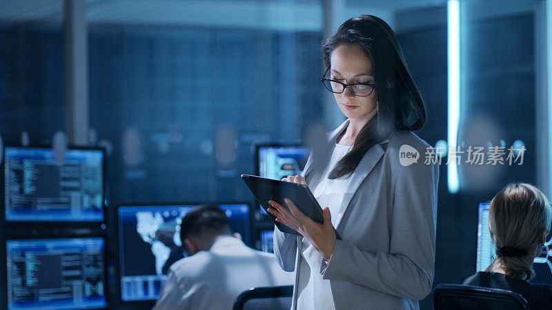 年轻女工程师在系统控制中心使用平板电脑。在后台，她的同事们在他们的工作空间里有许多显示有价值的数据的显示器。