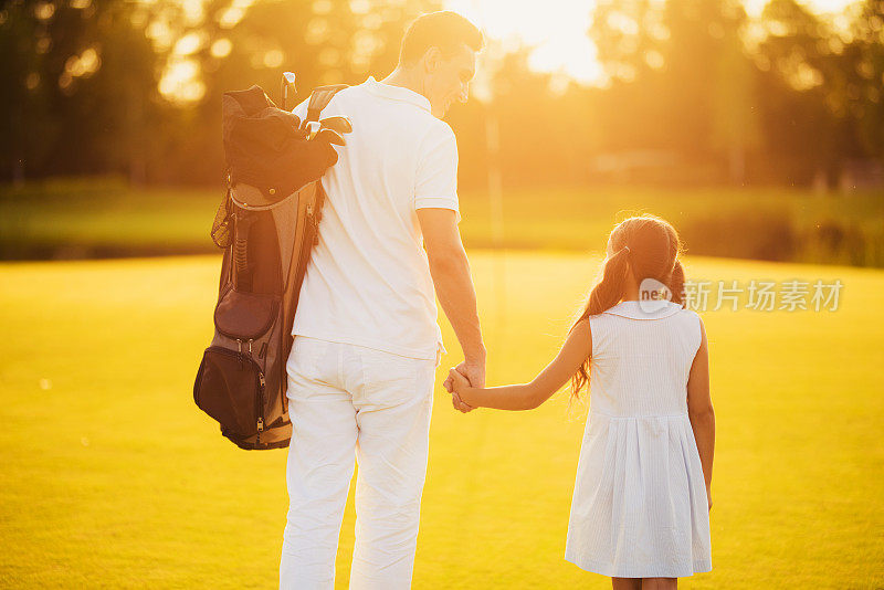 一个拿着高尔夫球杆包的男人和一个在夕阳背景下牵着手沿着高尔夫球场走的女孩