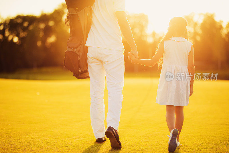 父亲和女儿在夕阳的背景下牵着手走在高尔夫球场上。