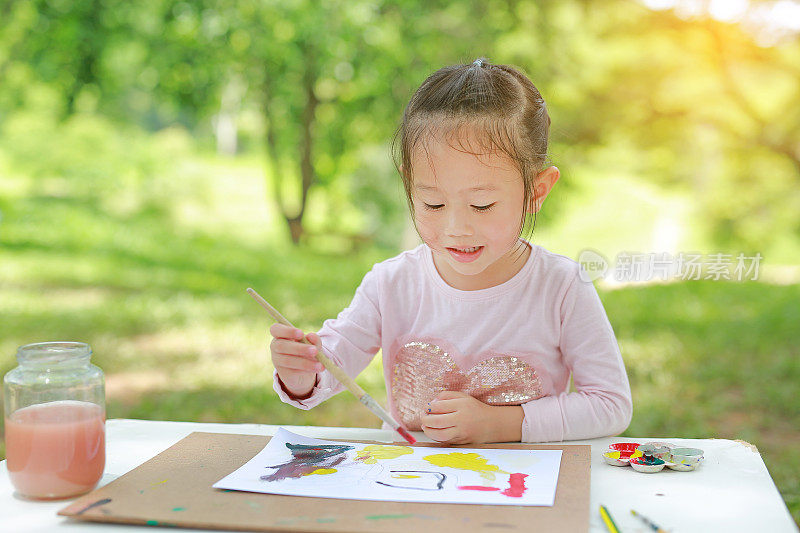 孩子坐在桌子上，在绿色的花园里画美术纸。