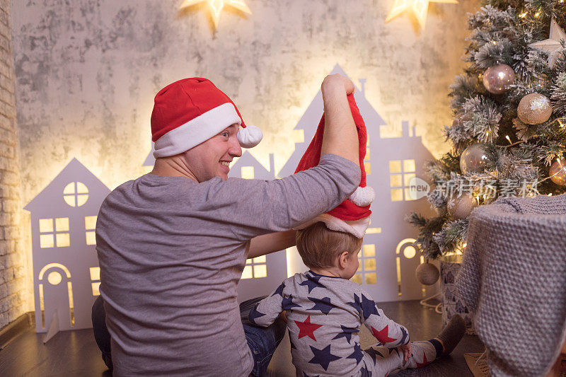 圣诞快乐和节日快乐的概念。爸爸和小罪恶戴着圣诞帽靠近圣诞树。圣诞前夜和家人在一起