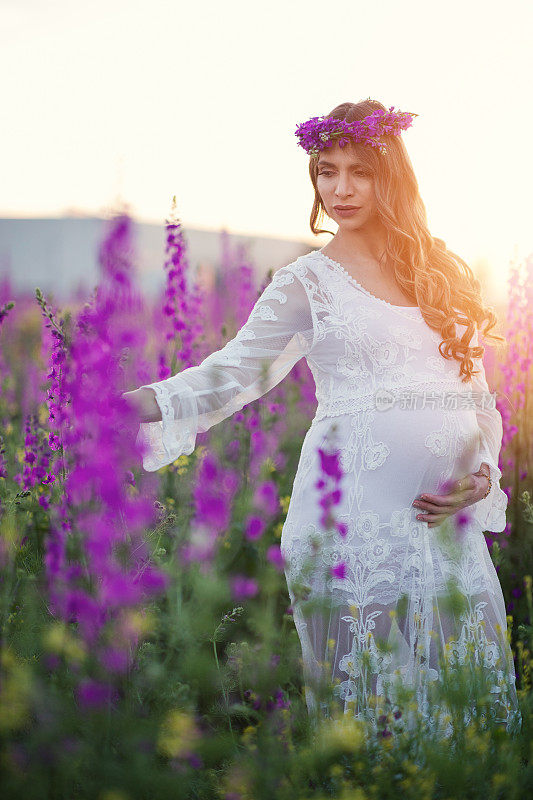 美丽的未来母亲，抚摸着自己的肚子，站在夕阳下的花丛中