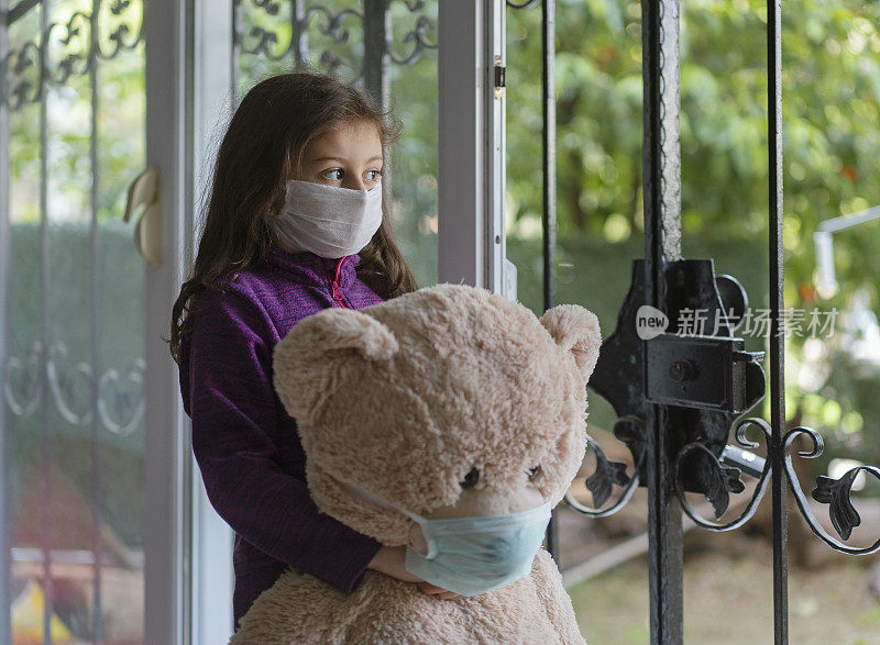 带着防护面具的可爱小女孩和她的泰迪熊站在窗边