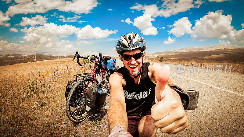 鼓舞人心的智能手机自拍肖像，兴奋快乐的白人男性骑自行车时竖起大拇指，看着相机的笑脸。骑自行车环游世界