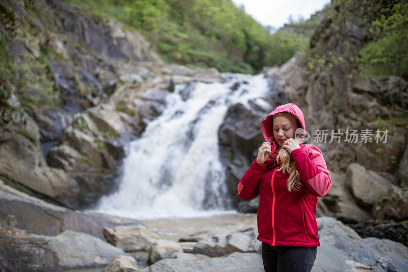 一位年轻的徒步女在瀑布旁整理她的雨衣