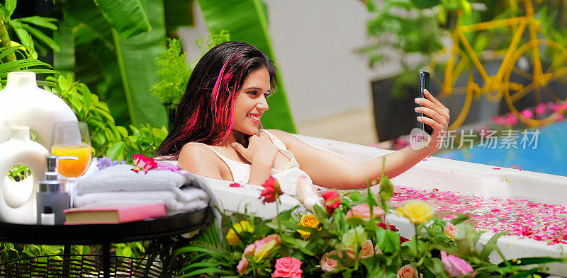 快乐的印度年轻成人放松女孩坐在浴缸新鲜的早晨沐浴在玫瑰花瓣的水享受暑假在酒店度假胜地。美丽微笑的z世代女子女士用手机远距离通话户外别墅之旅